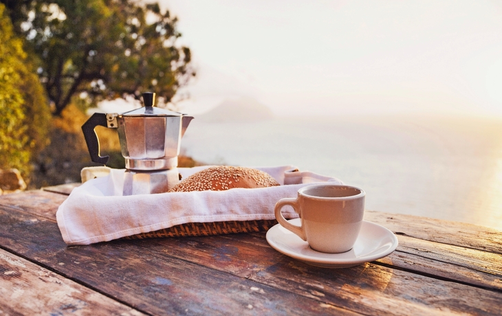 Mittelmeer-Frühstück, Kaffee und frischem Brot auf einem Tisch mit Bäumen und Blick auf das Meer im Hintergrund